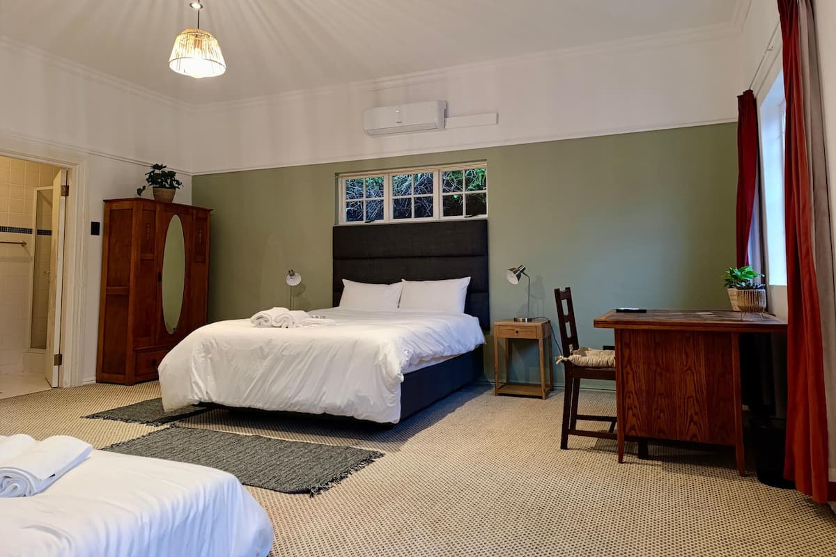 A hotel room with a large bed, a desk, a chair, a small table, a potted plant, a mirror, and a window with curtains.