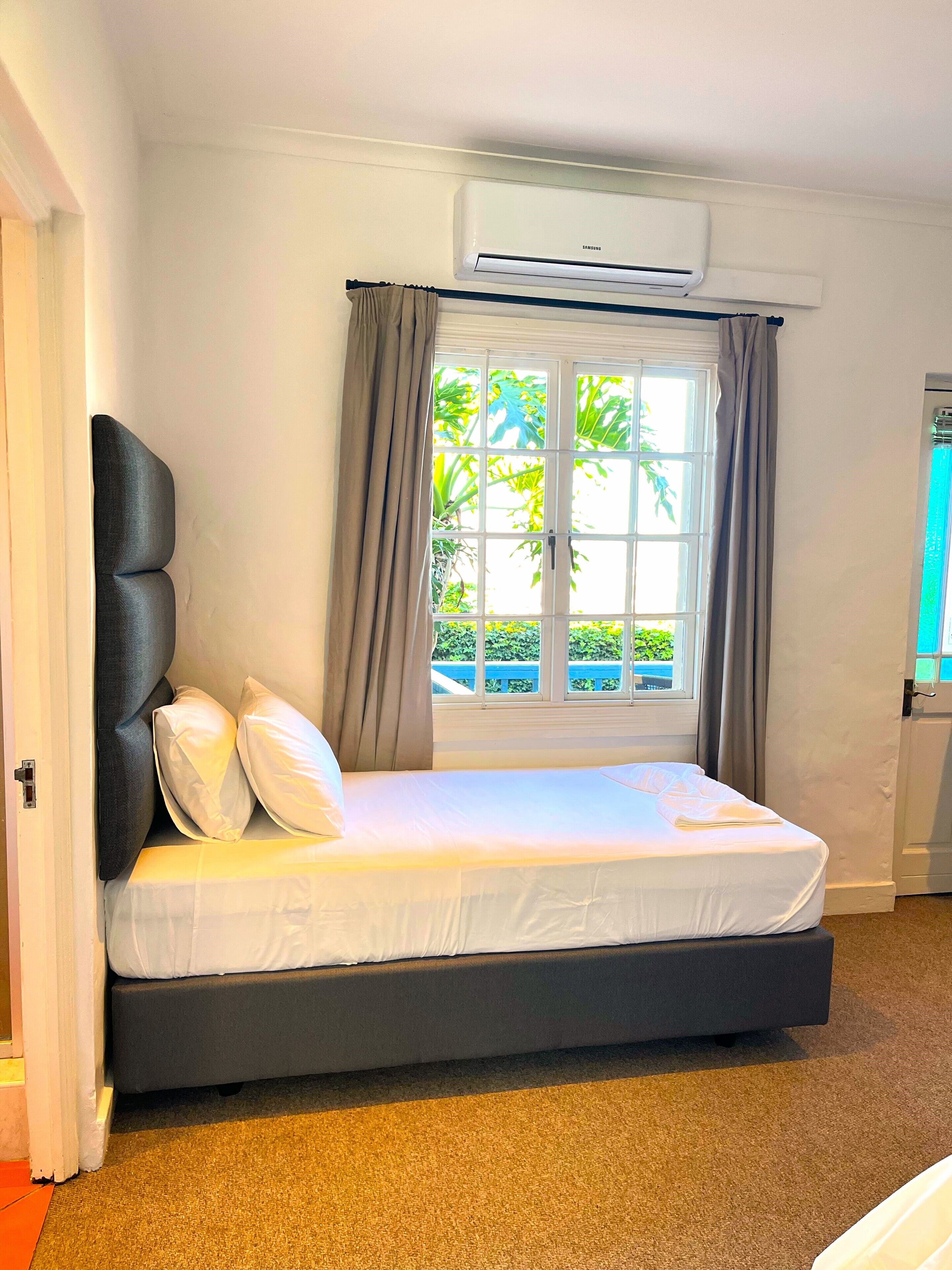 A bedroom with a bed, an air conditioning unit, a window with curtains, and a view of greenery outside.
