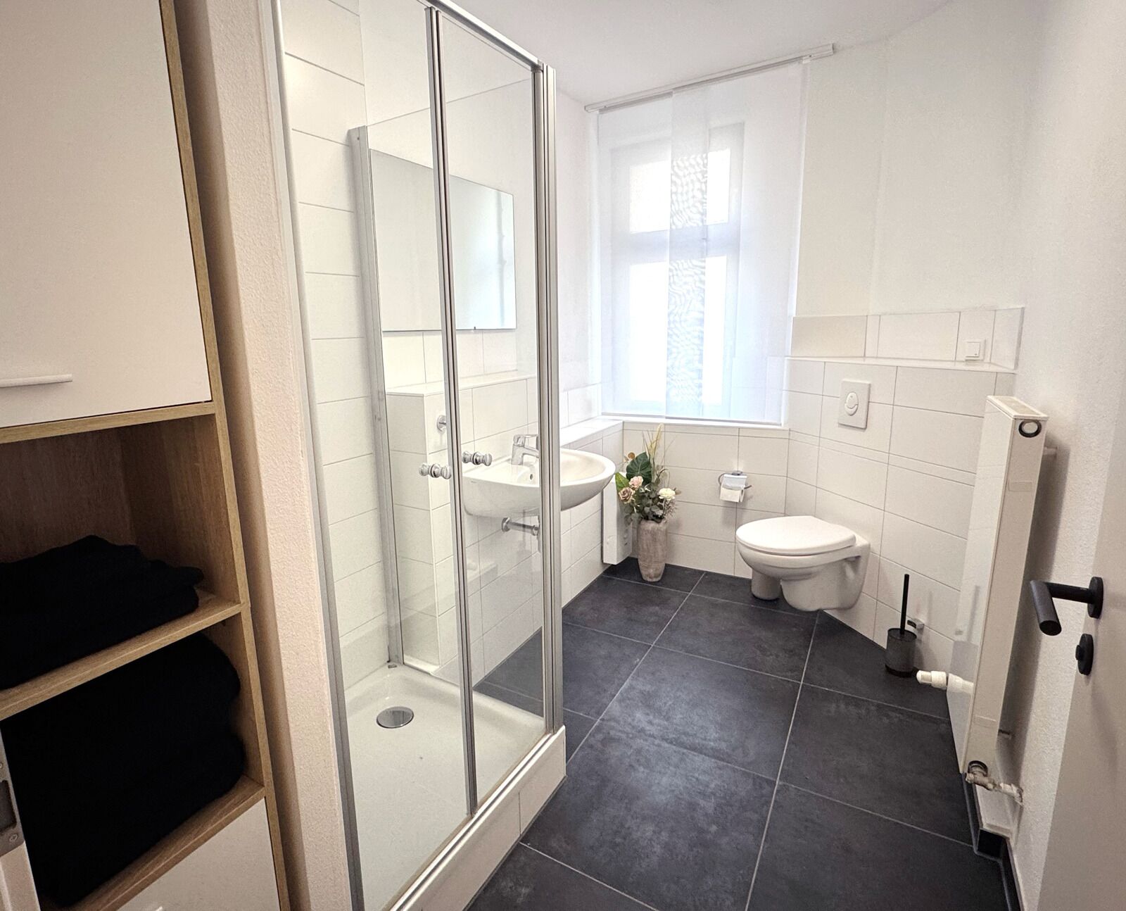 Ein modernes Badezimmer mit Duschkabine aus Glas, Waschbecken, Toilette und einem Fenster mit Jalousien.