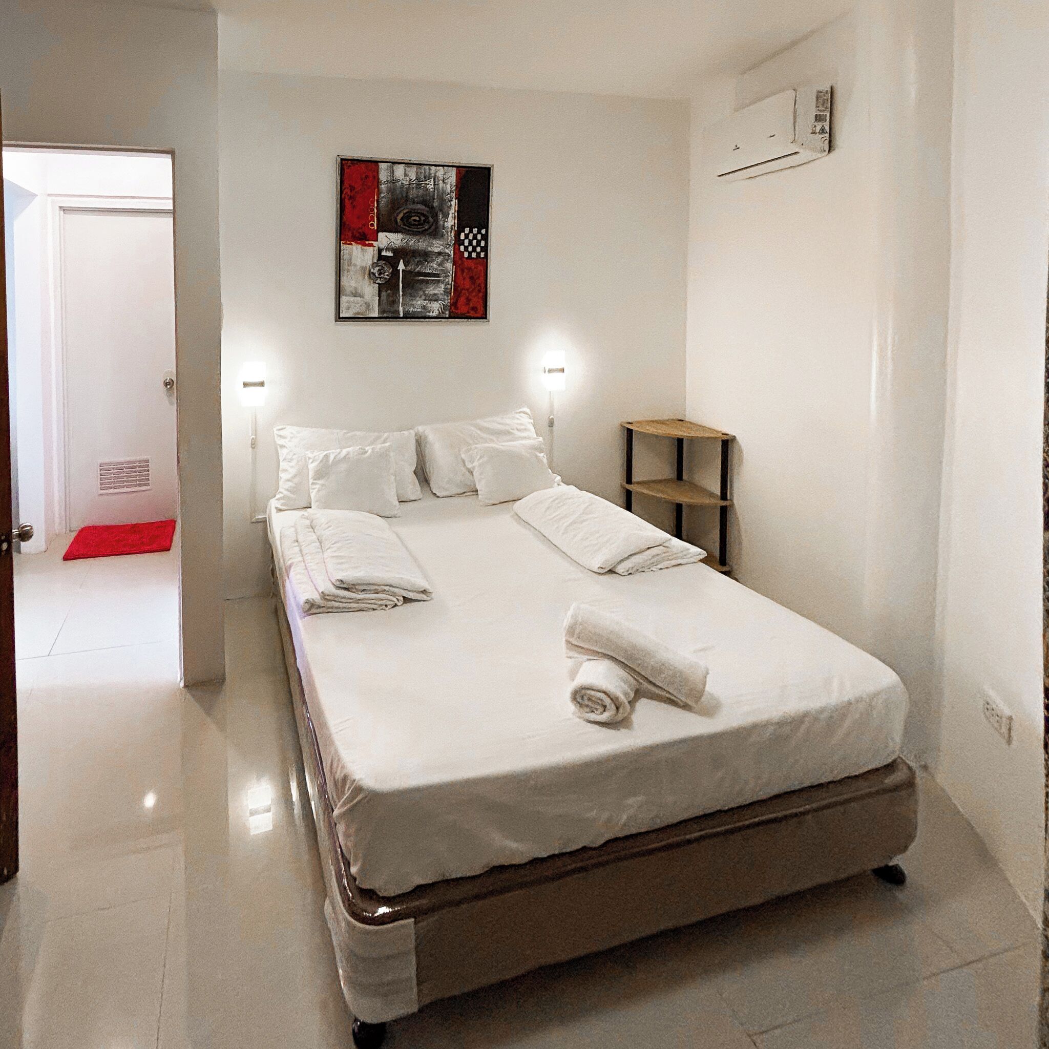 Un dormitorio con cama, almohadas, una mesita de noche, una obra de arte en la pared y una puerta que da a otra habitación.