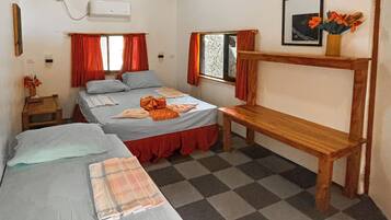 A room with two beds, a bench, a window with curtains, a framed picture, and a small table with a vase of flowers.