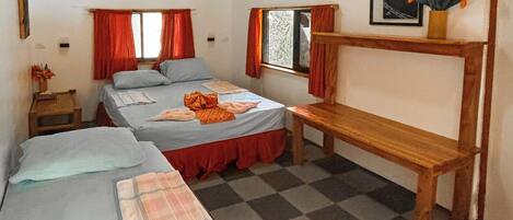 A room with two beds, a bench, a window with curtains, a framed picture, and a small table with a vase of flowers.