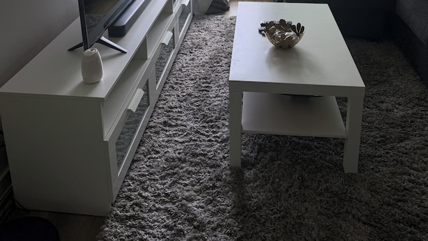 A modern living room with a flat-screen TV on a white stand, a white coffee table with a decorative bowl, a grey carpet, and a large window with a view of a green landscape and a road.