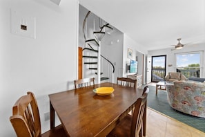 A dining area with a wooden table, chairs, a yellow bowl, a spiral staircase, a television, and a balcony with a view. - Bunk Bed Beach Base | 2BR Family Retreat at The Cove (Gulf Shores)