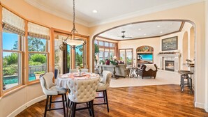 A spacious living room with a fireplace, a dining area with a round table and chairs, and large windows offering a view of a pool and greenery.
