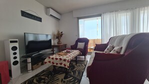 Ett vardagsrum med en platt-TV, en röd fåtölj, en soffa, ett soffbord och ett fönster med gardiner.