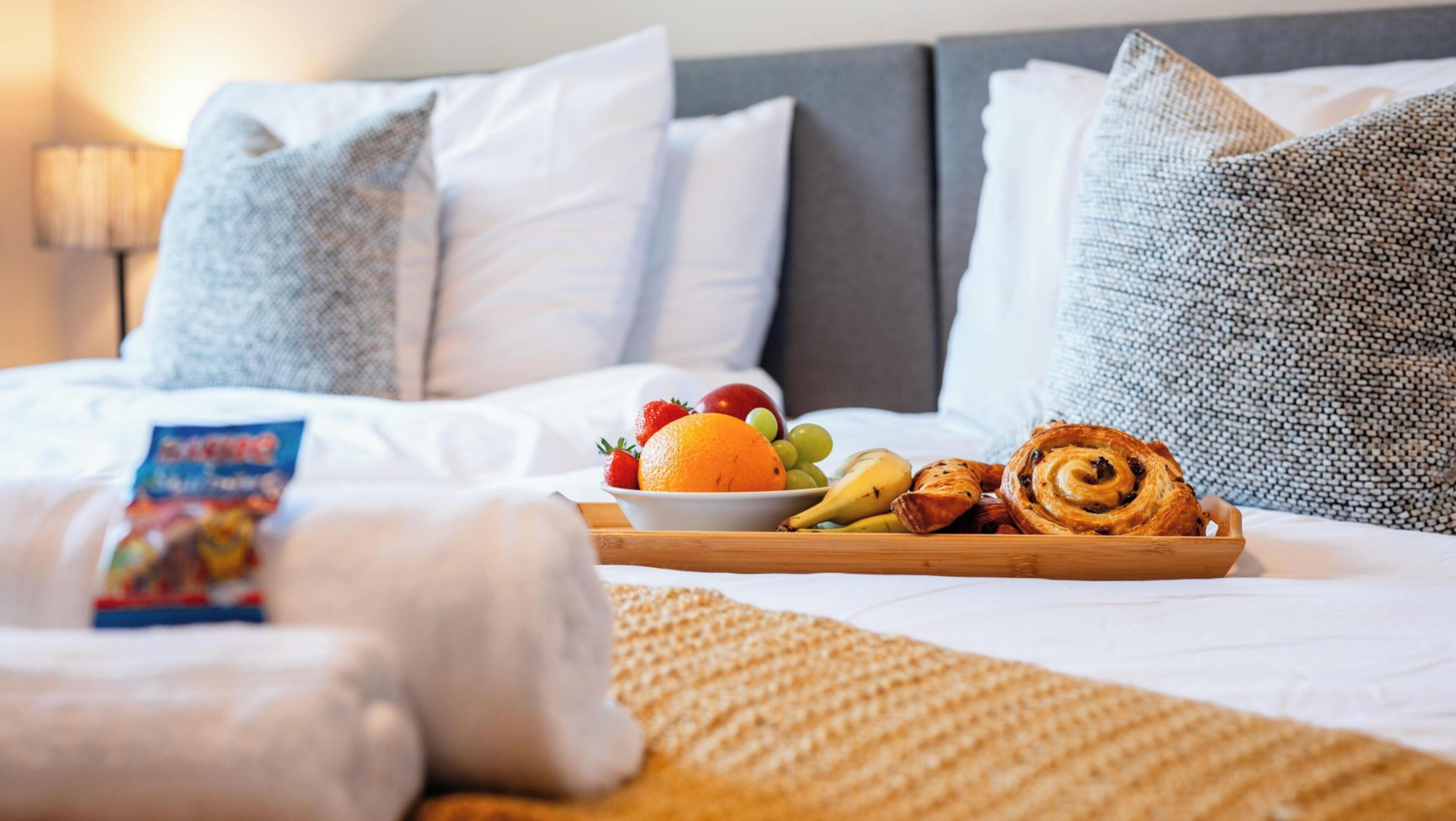 Een netjes opgemaakt bed met een dienblad vol ontbijtartikelen, zoals fruit en gebak, een opgerolde handdoek en een kussen.