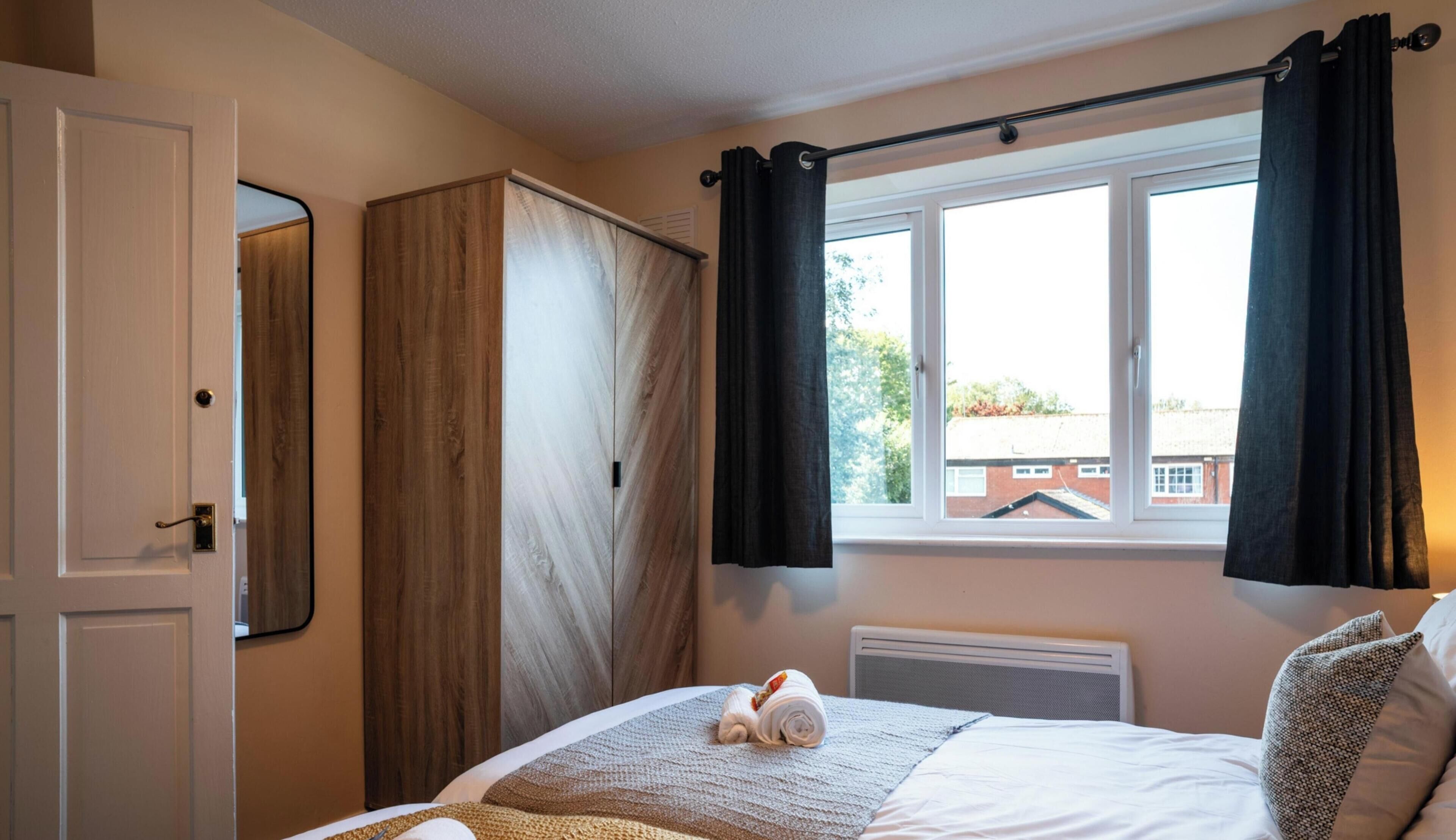 A hotel room with two beds, a large window with curtains, a radiator, and a mirror on the wall.