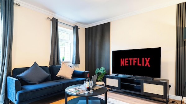 A modern living room with a sofa, a coffee table with a wine bottle and glasses, a TV with 'NETFLIX' on the screen, and a window with curtains.