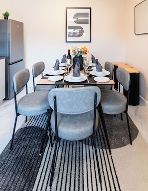 A modern dining area with a table set for six, grey upholstered chairs, a black and white striped rug, a refrigerator, and a framed abstract artwork on the wall.