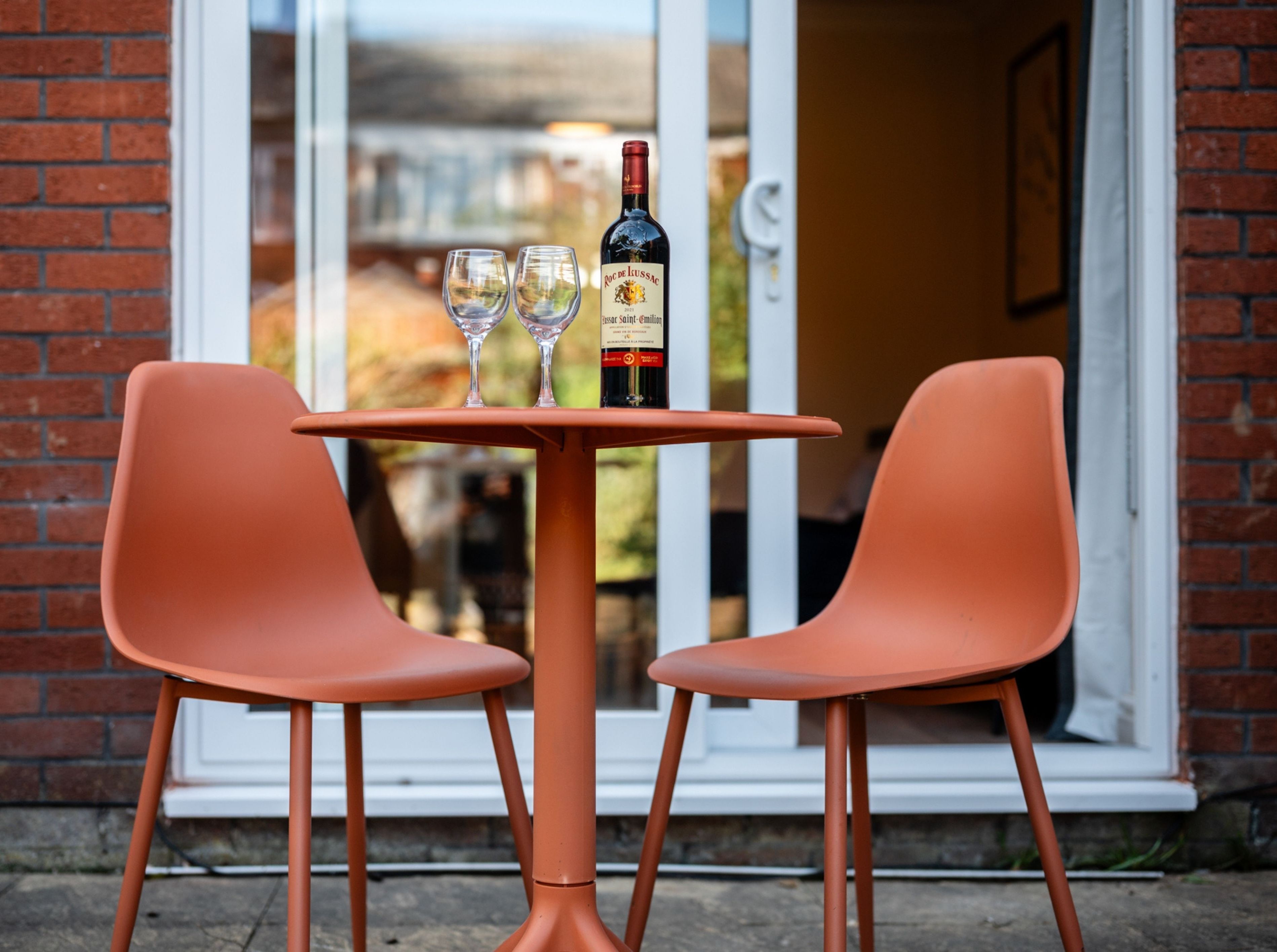 A table with two chairs and a bottle of wine, set against a brick wall with a glass door leading to a garden.