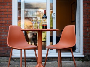 A table with two chairs and a bottle of wine, set against a brick wall with a glass door leading to a garden.