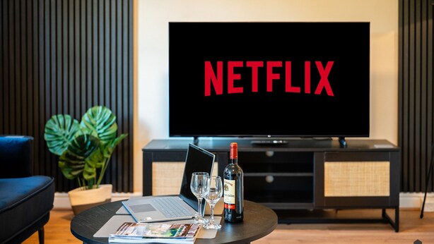 Moderni olohuone, jossa on suuri litteänäyttöinen televisio, jonka ruudulla näkyy Netflixin logo. Lisäksi huoneessa on kannettava tietokone, kaksi viinilasia ja viinipullo sohvapöydällä.