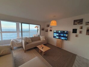 Ein Wohnzimmer mit einer Couch, einem Couchtisch, einem an der Wand montierten Fernseher und einem großen Fenster mit Meerblick.