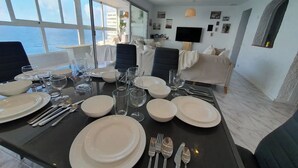Zona pranzo con tavolo imbandito per quattro persone, divano, televisore e vista sul mare.