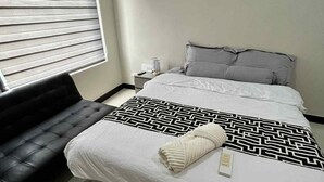 A hotel room with a bed, a black sofa, a nightstand with a lamp, and a window with blinds.