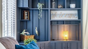 Ein dunkelblaues, eingebautes Regalsystem mit Dekorationsobjekten und einem 'Welcome'-Schild, ein gepolstertes Sofa mit einem blauen Kissen und eine beleuchtete Stehlampe.