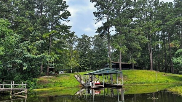 A wooden dock extending into a calm lake, surrounded by dense forest. A covered pavilion with steps is visible on the shore.