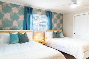 Habitación de hotel con dos camas individuales, una mesita de noche con lámpara, una ventana con persianas y una puerta.