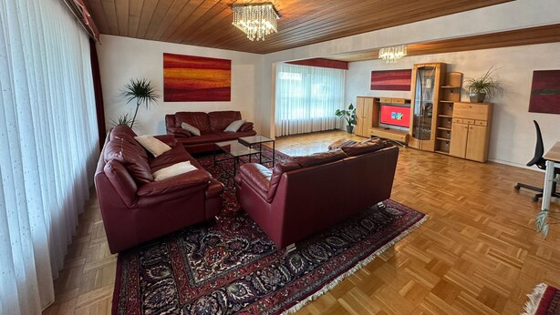 Ein geräumiges Wohnzimmer mit Holzboden, einem großen, roten Sofa, einem Couchtisch, einem Fernseher und einer Holzkommode mit Regalen.