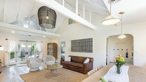 Een ruime woonkamer met een hoge plafond, een bruine leren bank, een glazen salontafel en een eettafel met een bloemistdecoratie.