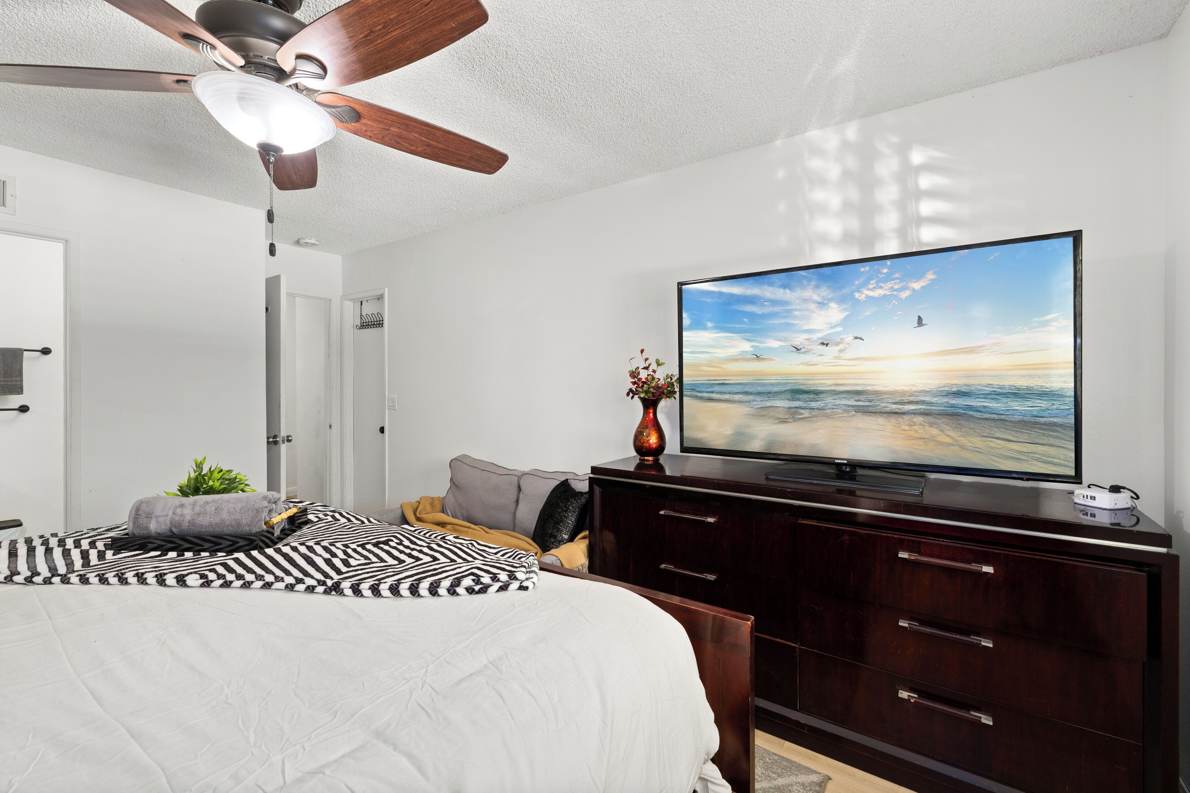 A bedroom with a bed, a ceiling fan, a TV on a dresser, and a view of the beach.
