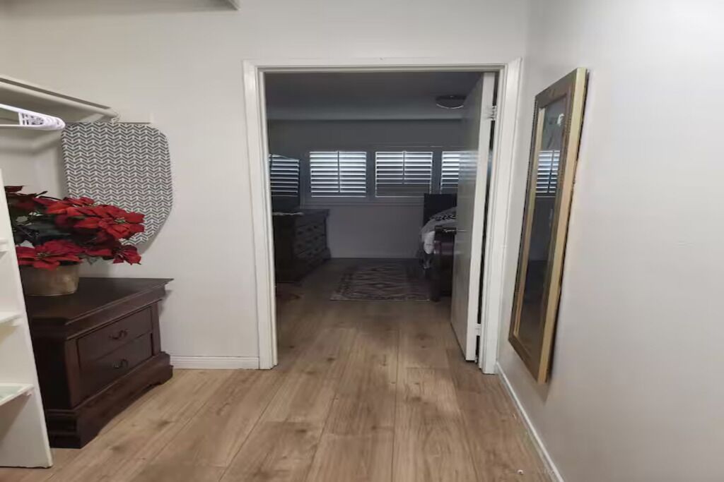 A narrow hallway with wooden flooring leading to a bedroom with a bed, a dresser, and a window with blinds.