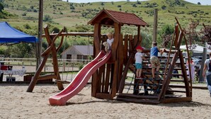 A playground with a slide and climbing structures, surrounded by a sandy area and a backdrop of green hills.