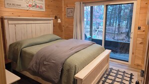 Ein Schlafzimmer in einer Holzhütte mit einem Bett, einem Nachttisch, einem Fenster mit Blick auf einen Wald und einem Schild, das eine Blumenwerkstatt bewirbt.