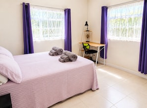 A bedroom with a bed, a desk, and two windows with purple curtains.