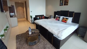 A modern hotel room with a large bed, a small side table, a water bottle, and a TV on the left wall. - High rise apartments (Gurugram)