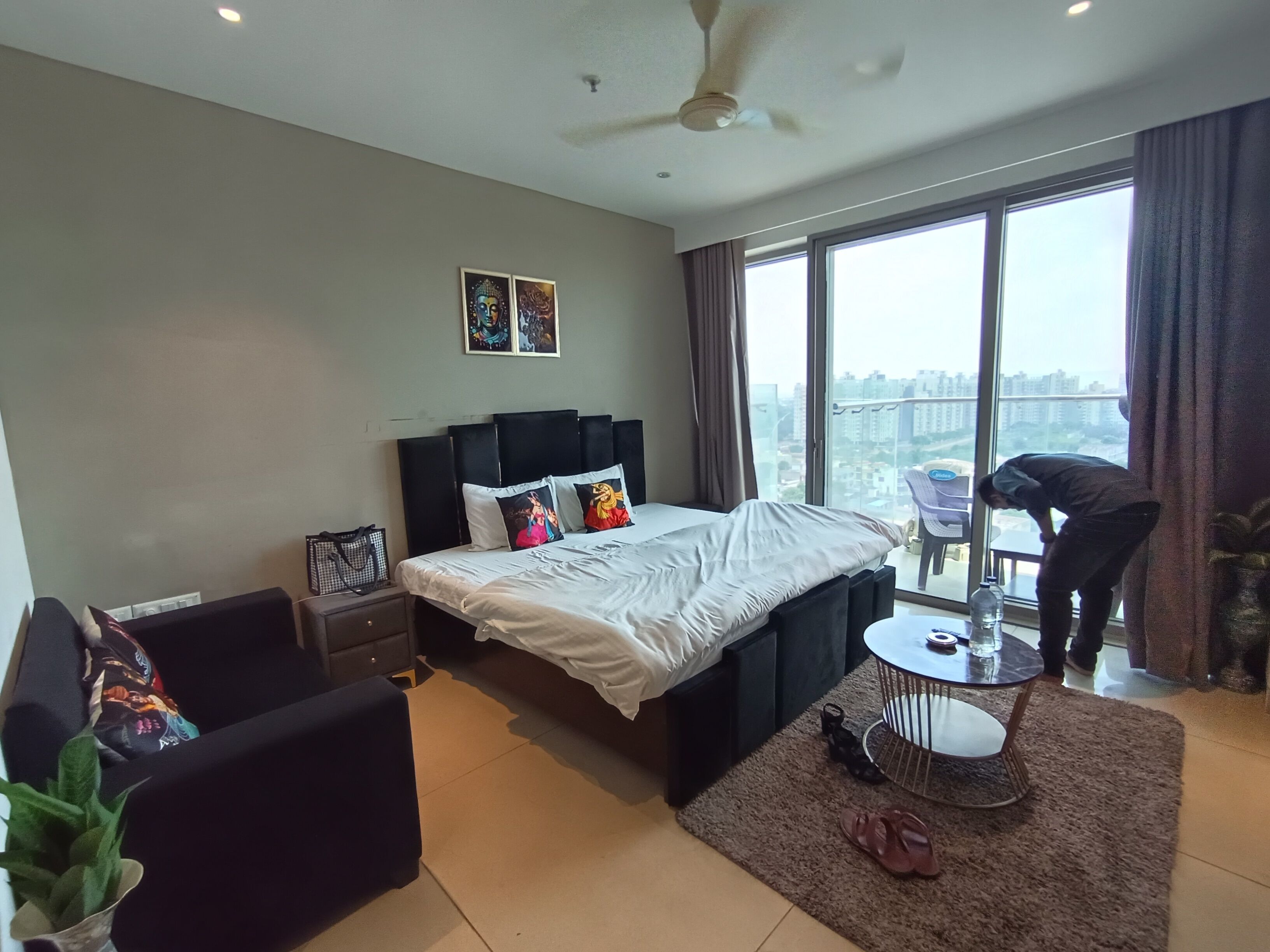 Een moderne hotelkamer met een groot bed, een klein tafeltje, een stoel, een kamerplant en uitzicht op de stad door grote ramen.