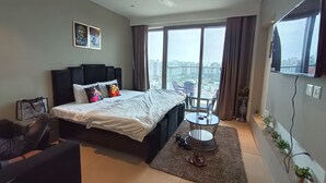 A modern hotel room with a large bed, a desk, a chair, a small table, and a balcony with a view of the city. - High rise apartments (Gurugram)