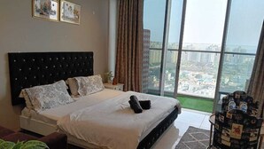A bedroom with a large bed, a window with a city view, and a decorative chair. - High rise apartments (Gurugram)