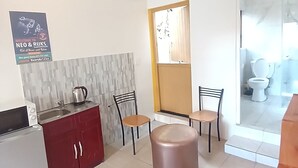 A compact kitchen with a microwave, sink, and cabinets. There are two chairs and a stool near the window. A poster on the wall advertises a guesthouse. - Neo & Ruks Guest House Parow (Cape Town)