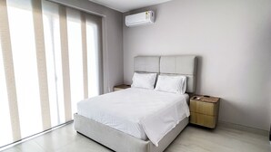 A modern bedroom with a large bed, a nightstand, a wall-mounted air conditioner, and a large window with blinds. - La Vie Es Belle Beach House (Santa Eleana)