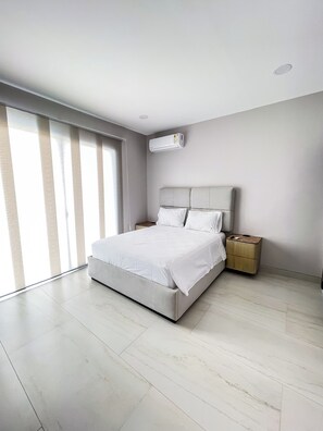 A modern bedroom with a large bed, a nightstand, a wall-mounted air conditioner, and a large window with blinds. - La Vie Es Belle Beach House (Santa Eleana)