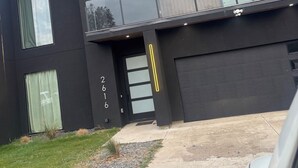 Une maison moderne à deux étages, dotée d’une façade sombre, de grandes fenêtres et d’un balcon avec des garde-corps en verre.