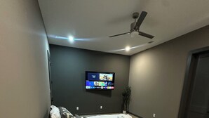 Une chambre à coucher moderne avec un lit, un téléviseur fixé au mur, un ventilateur de plafond et une table de chevet.