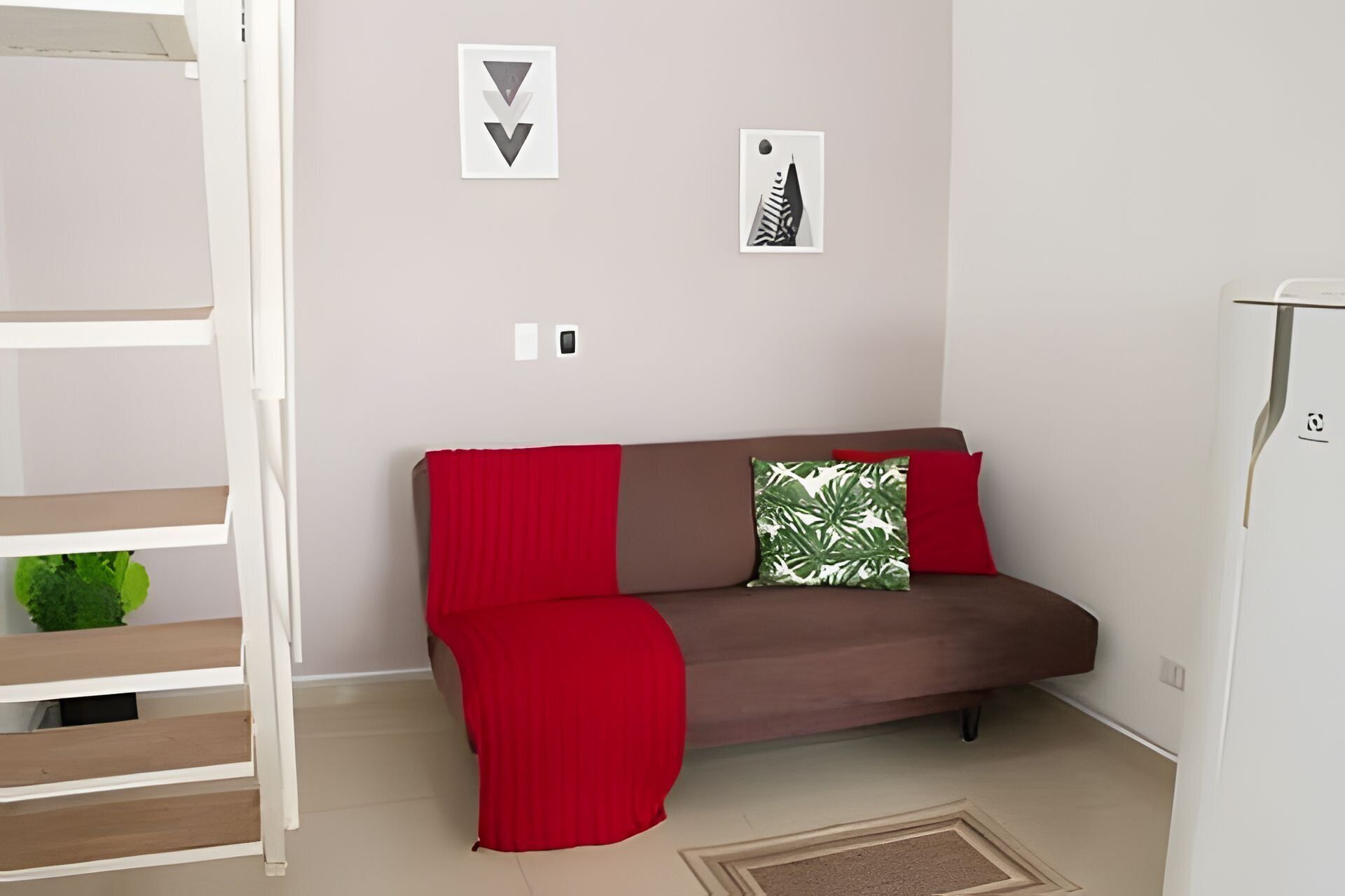 Un salon moderne avec un canapé marron, un plaid rouge et un coussin vert. Deux tableaux encadrés sont accrochés au mur, et il y a un réfrigérateur blanc.