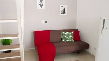 Un salon moderne avec un canapé marron, un plaid rouge et un coussin vert. Deux tableaux encadrés sont accrochés au mur, et il y a un réfrigérateur blanc.