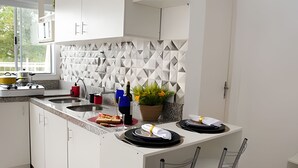 Uma cozinha moderna com armários brancos, bancada de mármore e painel geométrico. Há uma pequena ilha com uma pia, fogão e um vaso de planta.