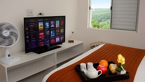 Quarto de hotel com televisão sobre um suporte branco, um ventilador, uma cama com cabeceira marrom e uma janela com vista para vegetação.