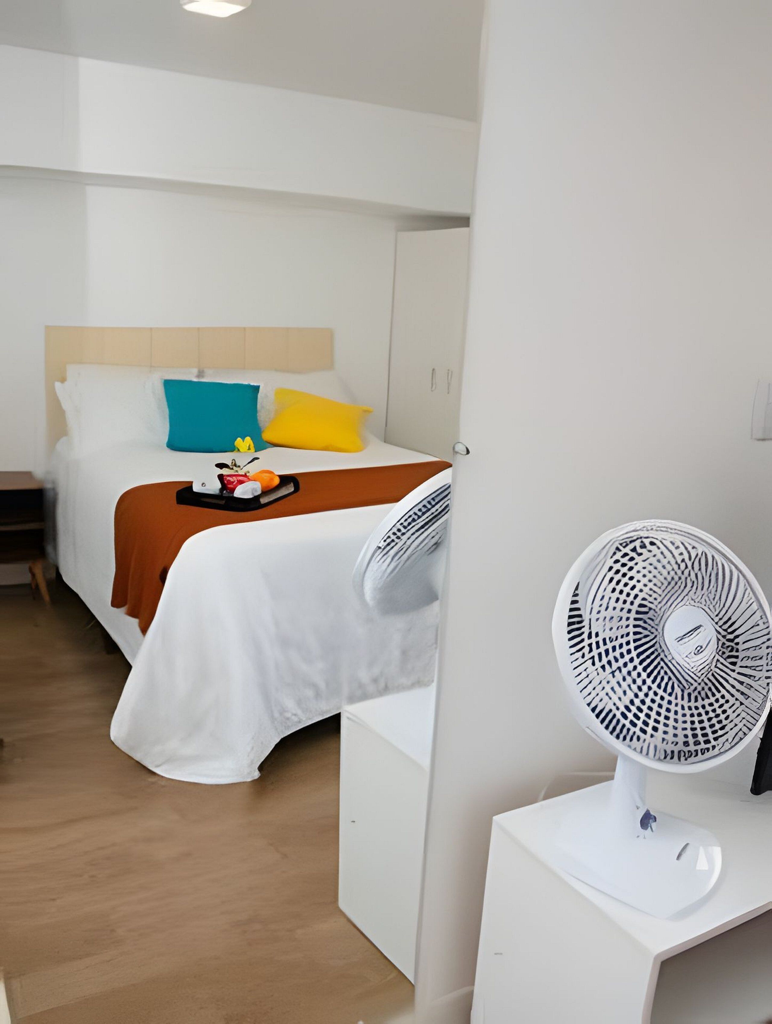 Habitación de hotel con cama, mesita de noche, ventilador blanco y suelo de madera.