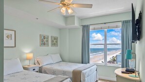 Ein Zimmer mit zwei Betten, einem Deckenventilator, einem Fenster mit Blick auf den Strand und einem Schreibtisch mit Lampe.
