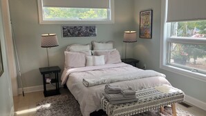 A bedroom with a bed, two nightstands, two wall-mounted lamps, a window with a view, and a framed picture on the wall.