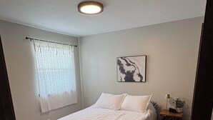 Une chambre à coucher avec un lit, une table de chevet, une lampe, une fenêtre avec des rideaux et un luminaire au plafond.