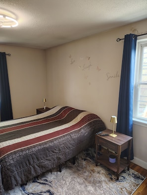 A bedroom with a bed, a bedside table with a lamp, a wall with decorative words, and a window with curtains.