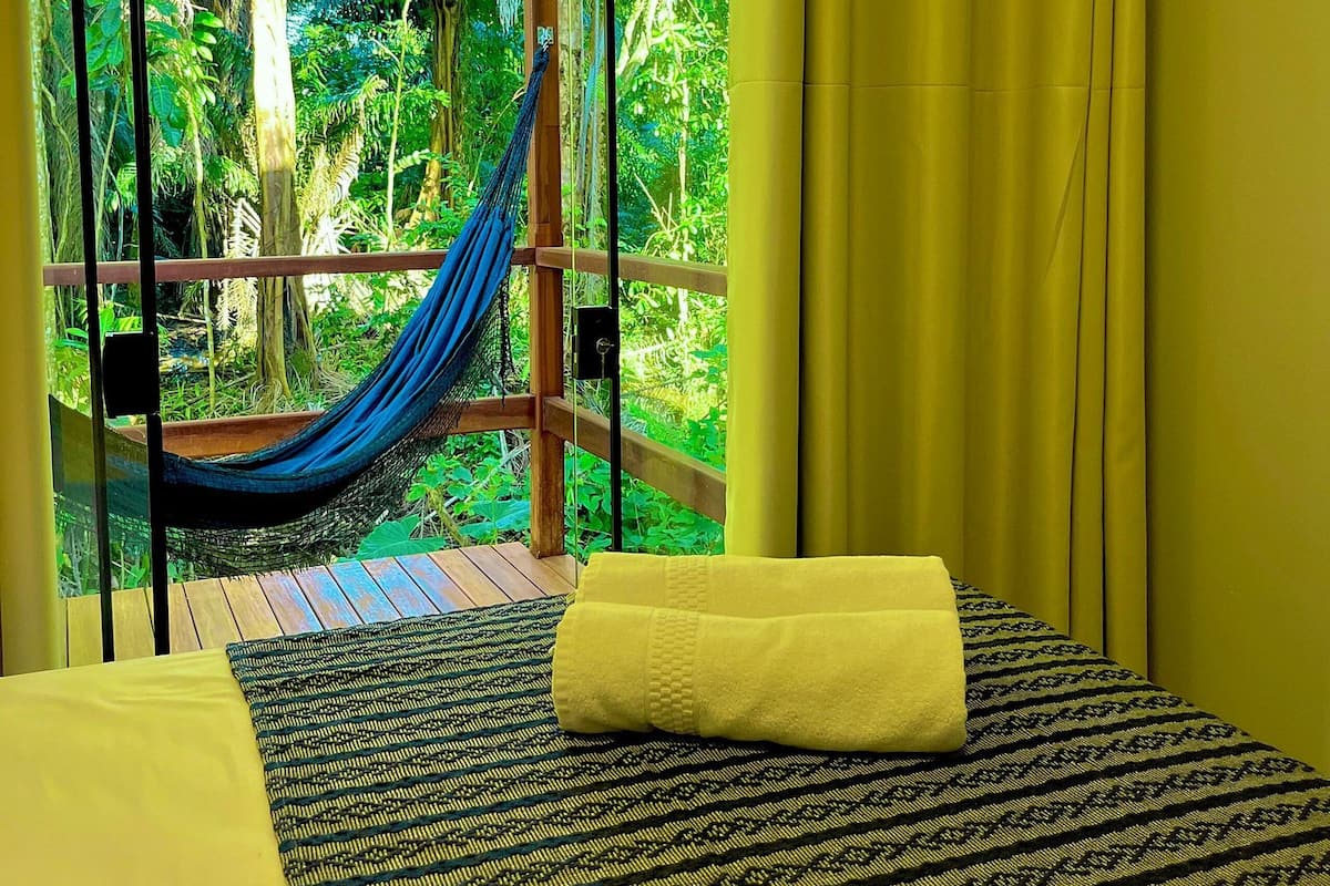 Une chambre avec un lit, un hamac et une vue sur un jardin luxuriant, visible à travers de grandes fenêtres.