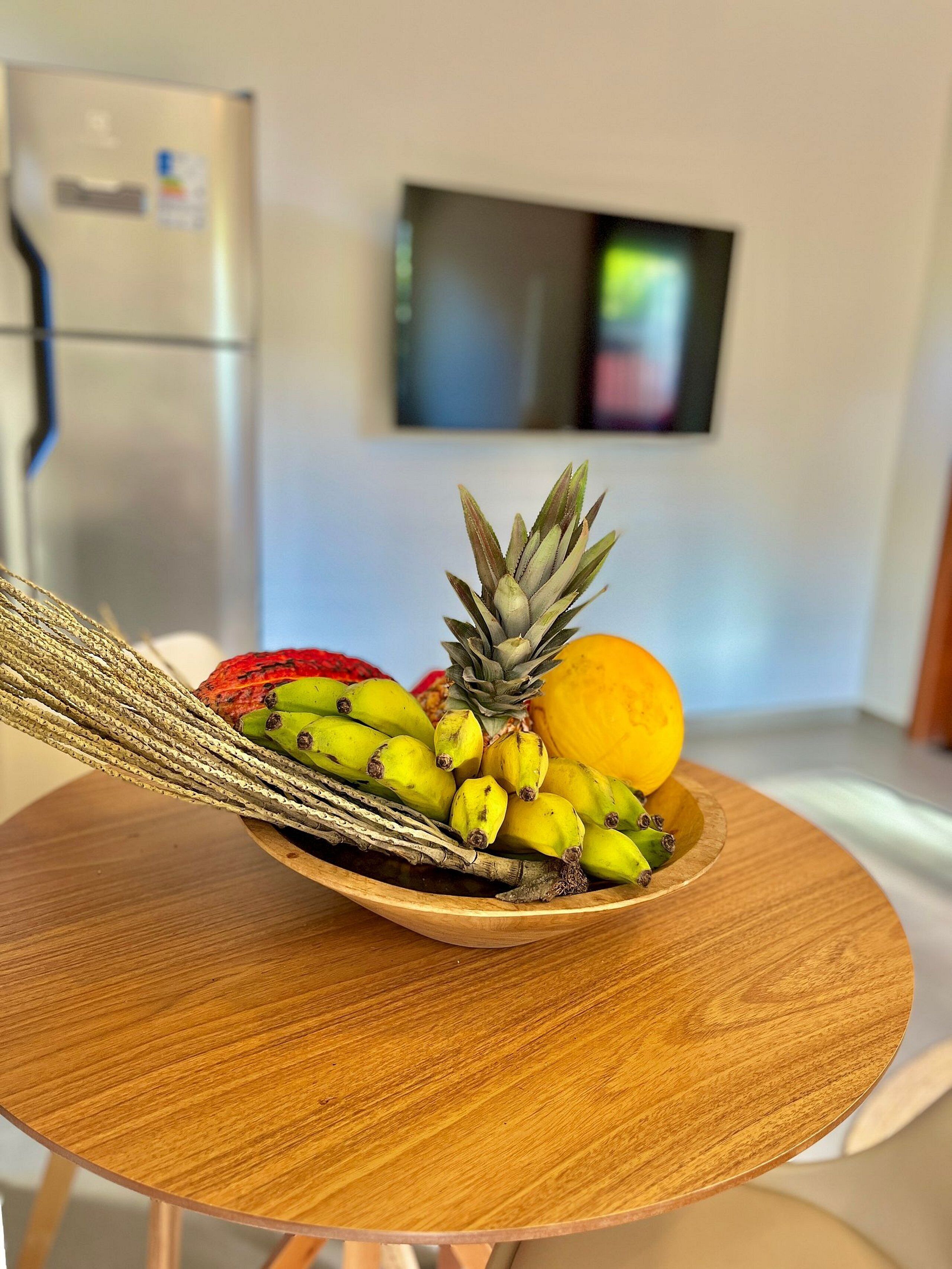 A wooden bowl on a round table with a variety of fruits including bananas, a pineapple, and a mango.